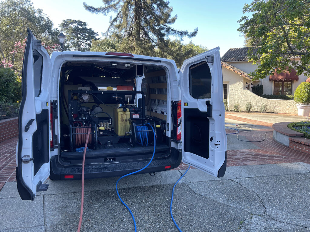 A utility van with open doors showing organized equipment and tools, parked near a residential building with hoses leading out.